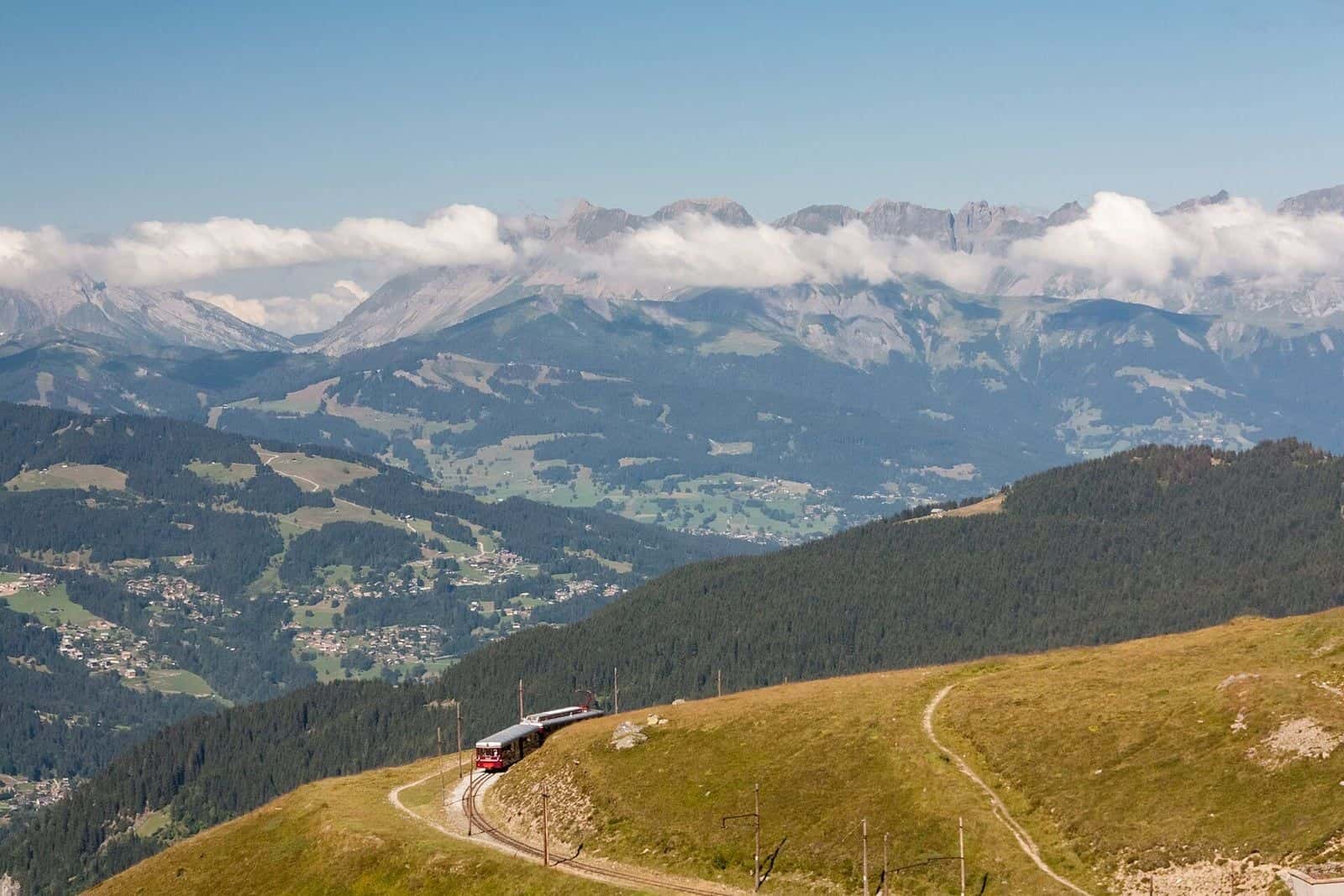 Mont Blanc Tramway train climbing through the pastures above Saint-Gervais, snowy peaks and a sea of clouds in the distance